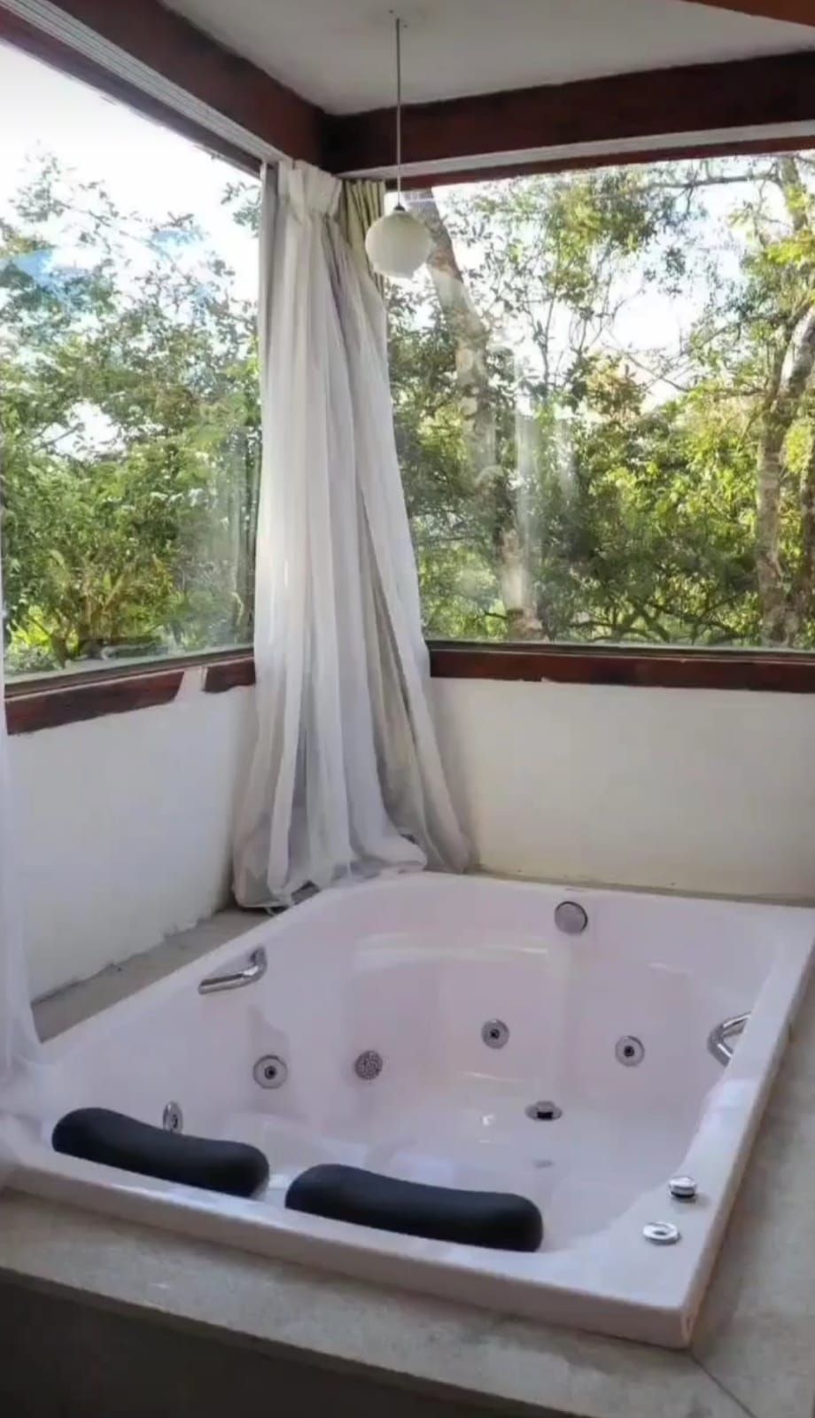 Uma banheira de hidromassagem branca com dois encostos de cabeça pretos, posicionada em um quarto com grandes janelas com vista para uma exuberante floresta verde.