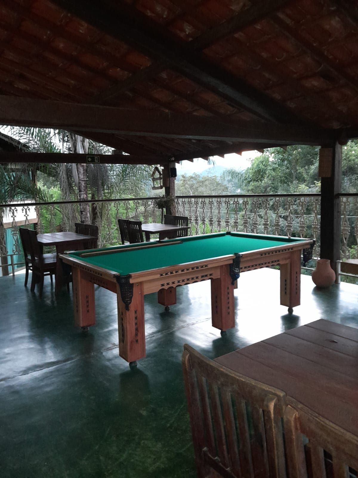 Uma mesa de bilhar verde está localizada em uma varanda externa coberta, com cadeiras de madeira e uma vista panorâmica das árvores ao fundo.