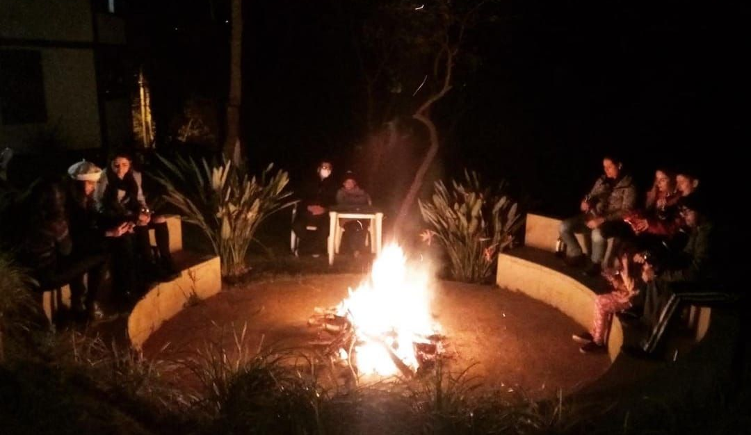 Um grupo de pessoas está sentado ao redor de uma fogueira bem iluminada em uma área circular ao ar livre, à noite.