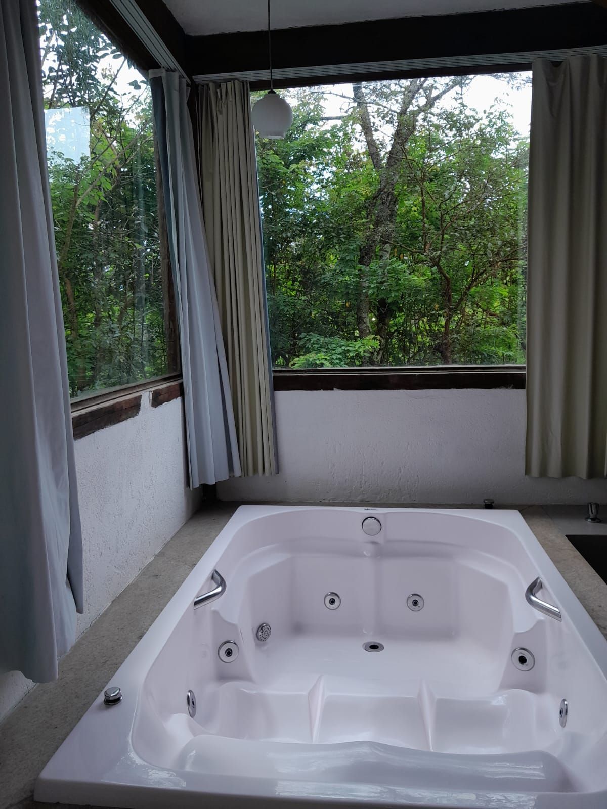 Uma banheira de hidromassagem branca está localizada em um quarto com grandes janelas que oferecem vista para uma floresta verdejante e exuberante, emoldurada por cortinas leves.