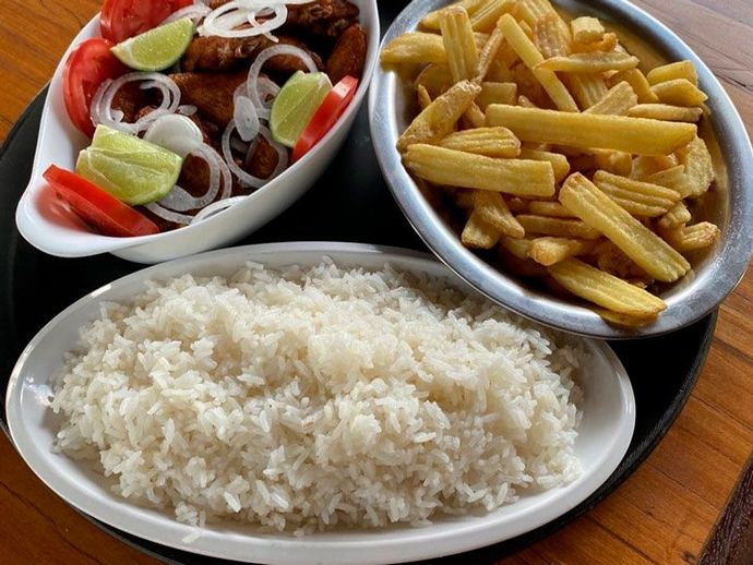 Uma refeição composta por arroz branco, batatas fritas e um prato coberto com rodelas de tomate, anéis de cebola e gomos de limão, servido numa bandeja preta.