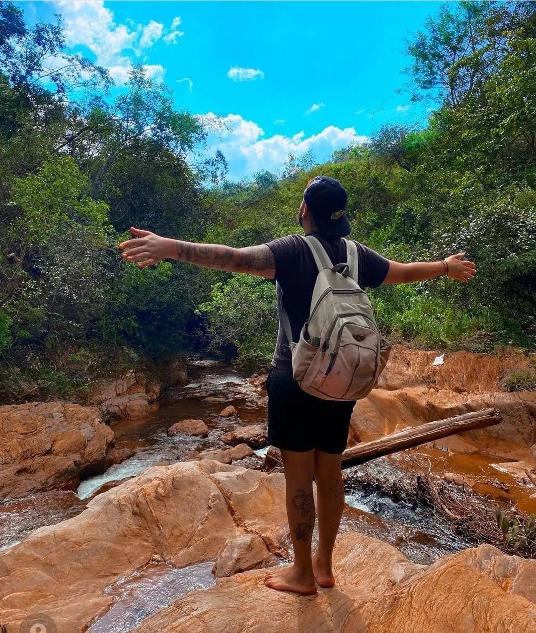 Uma pessoa com uma mochila está descalça sobre pedras à beira de um riacho, com os braços estendidos, de frente para uma floresta exuberante.
