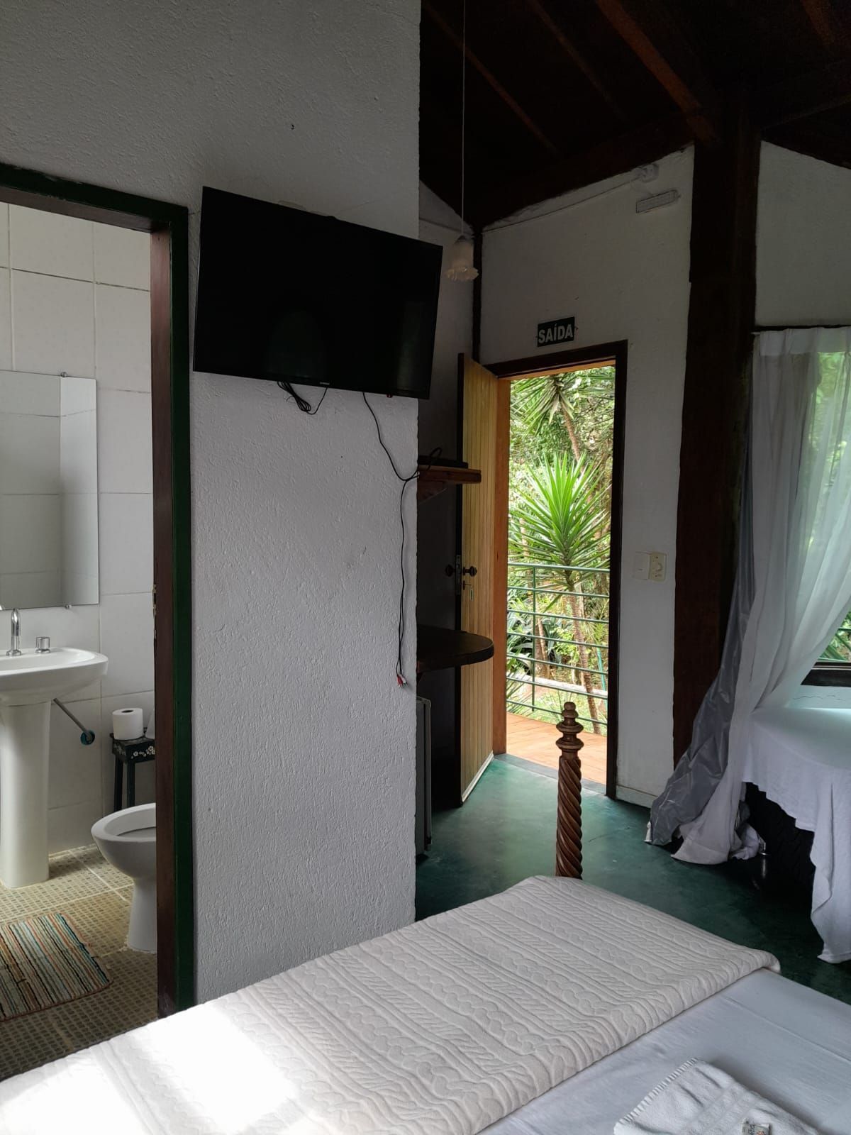 Um quarto com uma TV montada na parede, uma cama em primeiro plano, a porta do banheiro aberta e uma saída que leva à natureza.