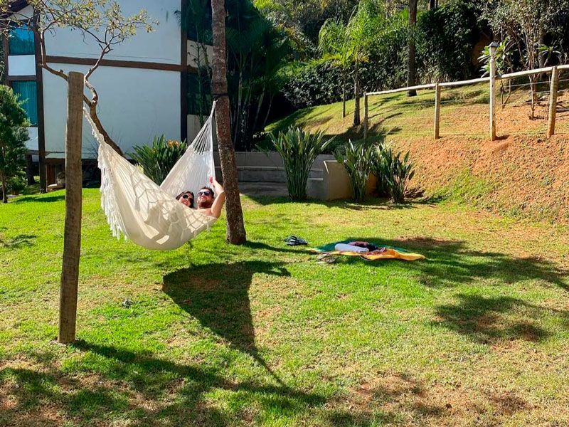 Duas pessoas relaxando em uma rede branca estendida entre árvores em um quintal ensolarado e gramado.