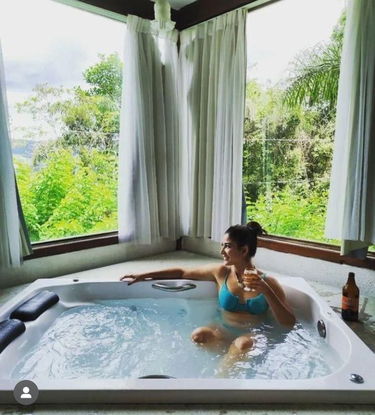 Uma pessoa de biquíni azul relaxa em uma banheira de hidromassagem com uma bebida, observando a vegetação exuberante através de grandes janelas de vidro.