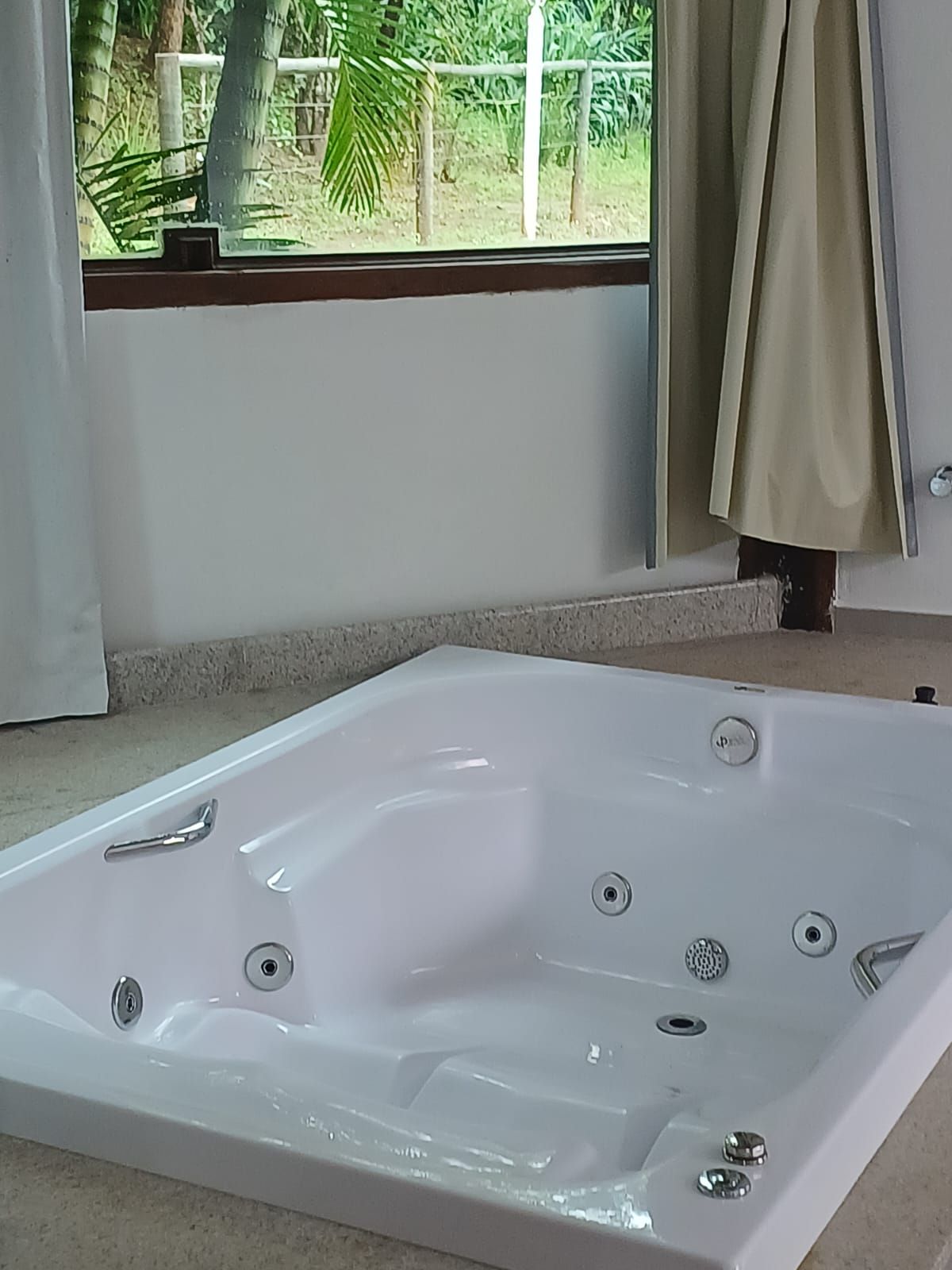 Uma banheira de hidromassagem branca fica ao lado de uma grande janela com cortinas bege, com vista para uma exuberante área verde externa.