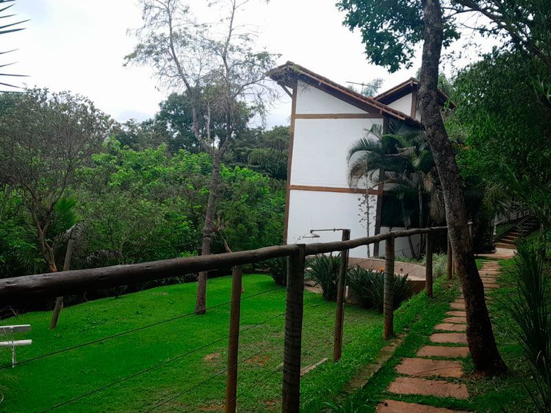 Uma casa branca de dois andares com estrutura de madeira, aninhada entre árvores, ao lado de um gramado e um caminho de pedra.