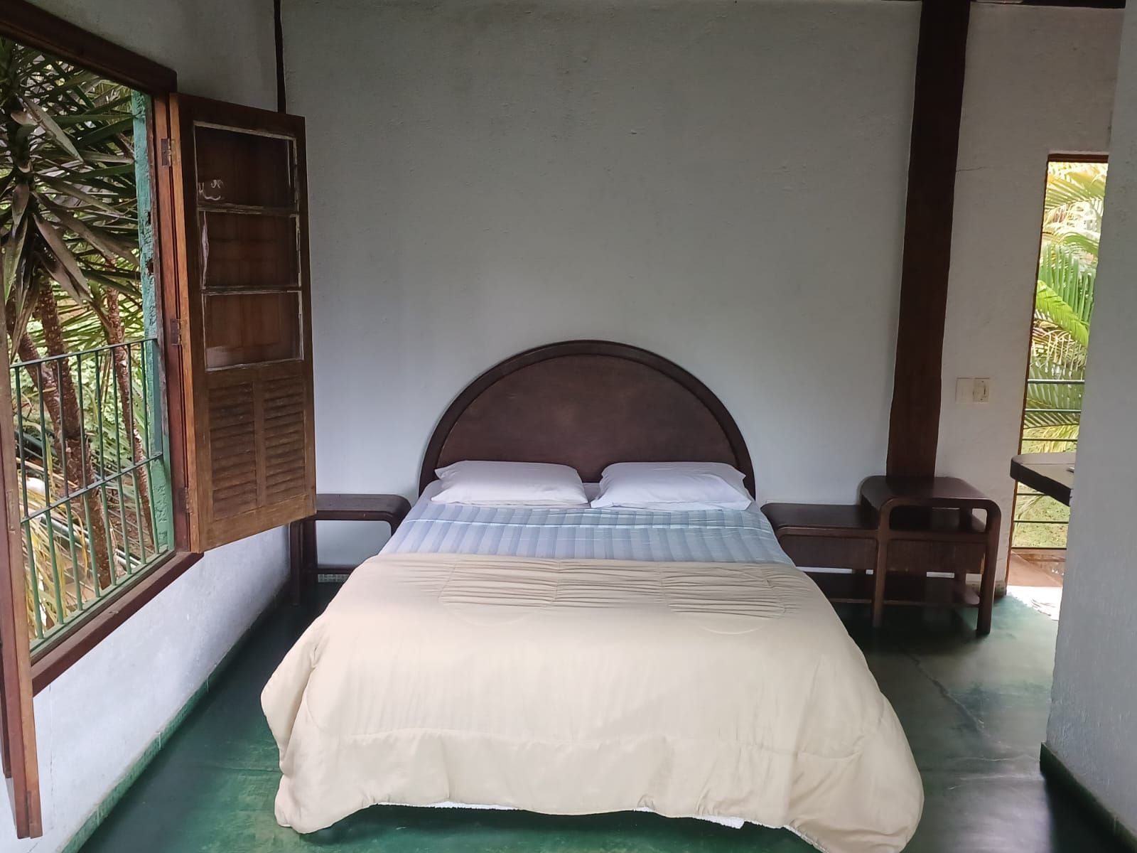 Um quarto com uma cama de casal, duas mesas de cabeceira de madeira e uma grande janela com vista para as árvores, com piso verde claro.