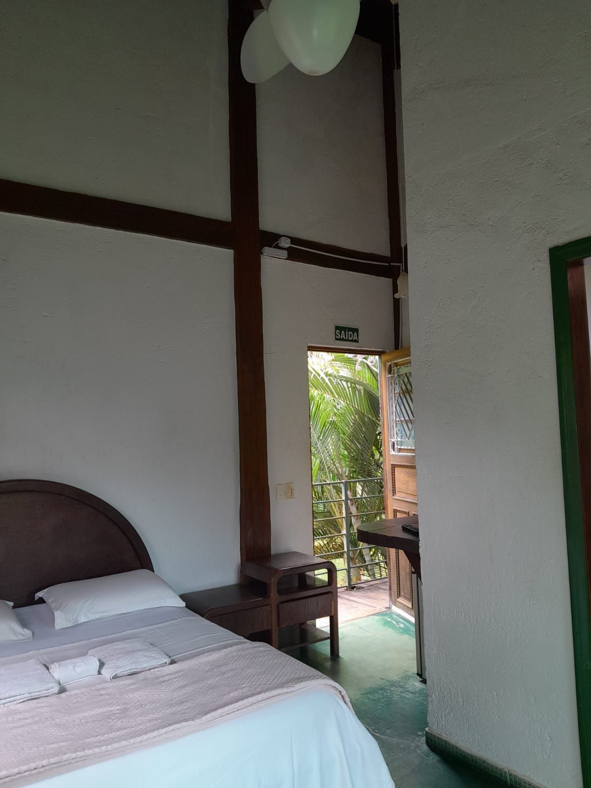 Um quarto com uma cama e uma parede com moldura de madeira, que se abre para uma varanda com plantas verdes sob um céu claro.