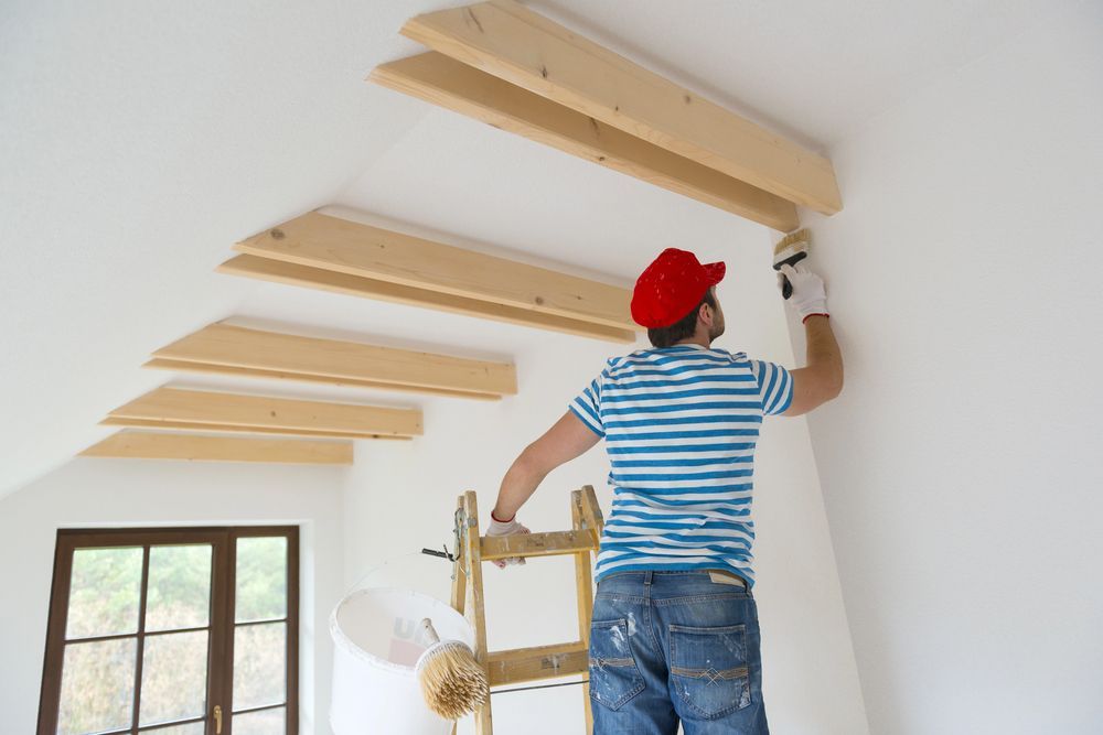 Painter on ladder painting white wall near wood beams.