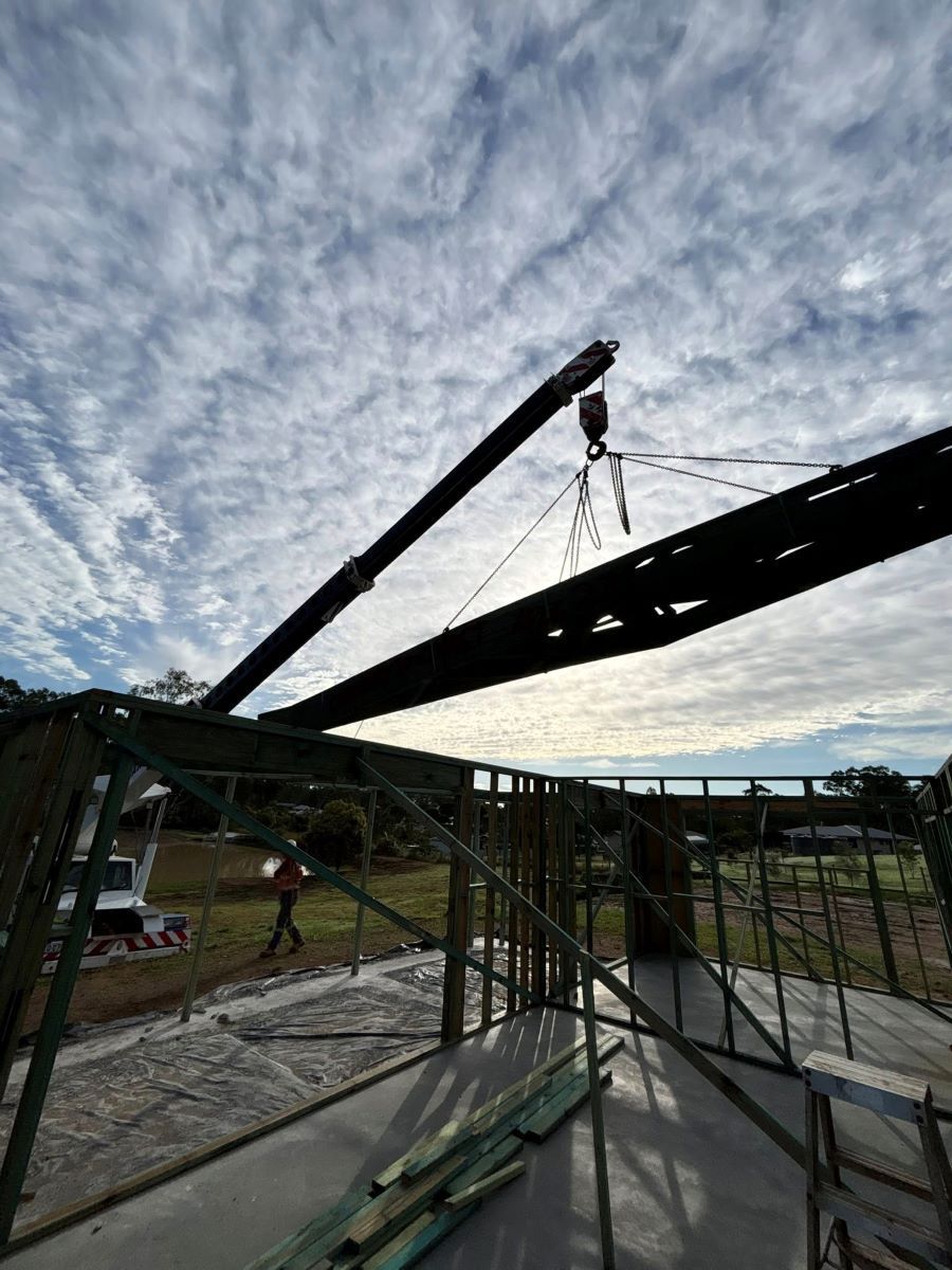 A Crane Lifts a Long, Dark Beam During the Construction of A Building — Country to Coast Homes In Bundaberg, QLD