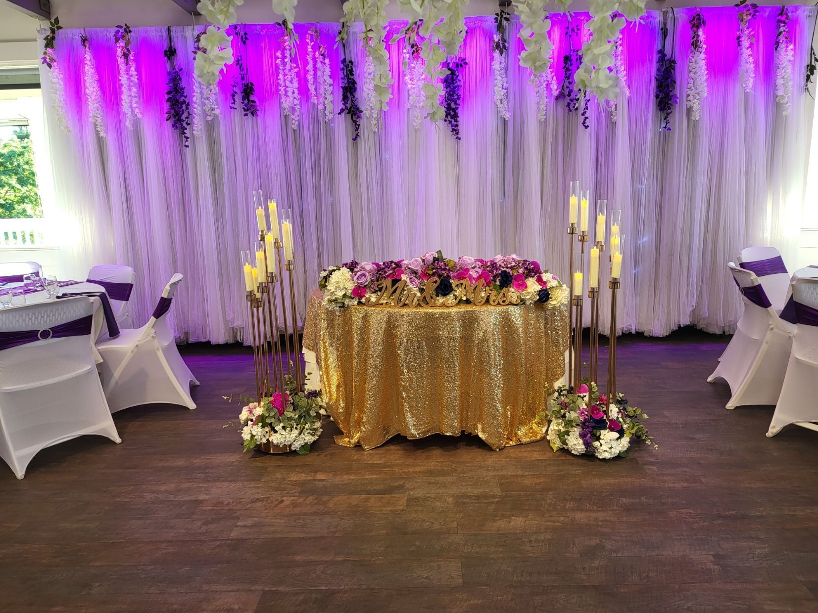 Gold-draped table with pink flowers and white curtains under purple lighting in an event venue
