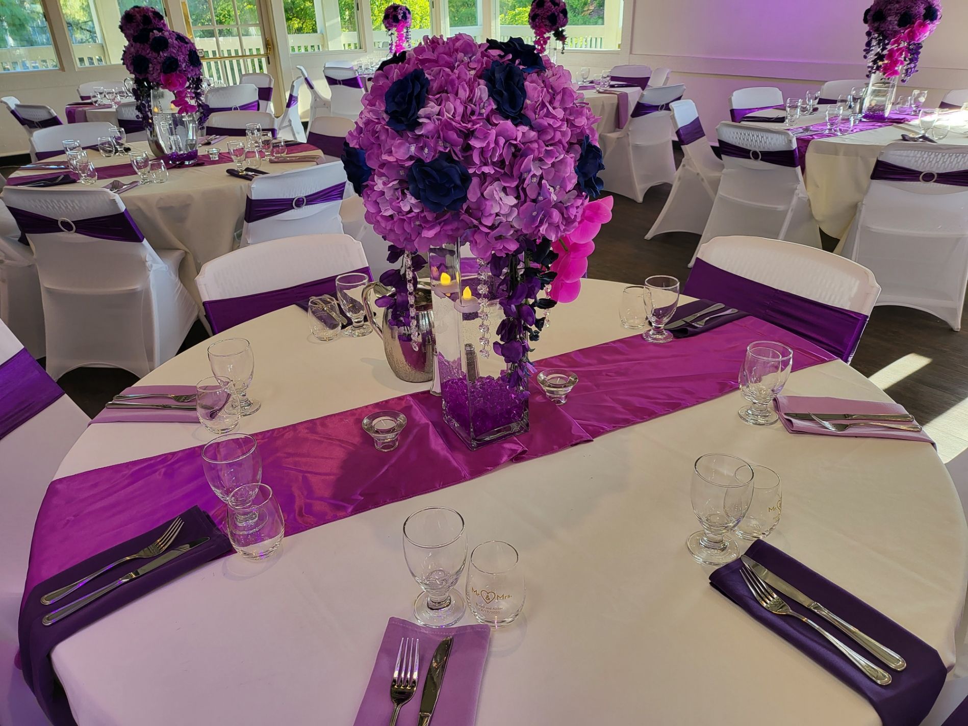 Purple floral centerpieces on decorated banquet tables with white chairs and silver place settings
