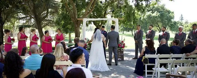 Outdoor wedding ceremony with bride and groom at the altar, bridesmaids in pink, and guests seated on white chairs