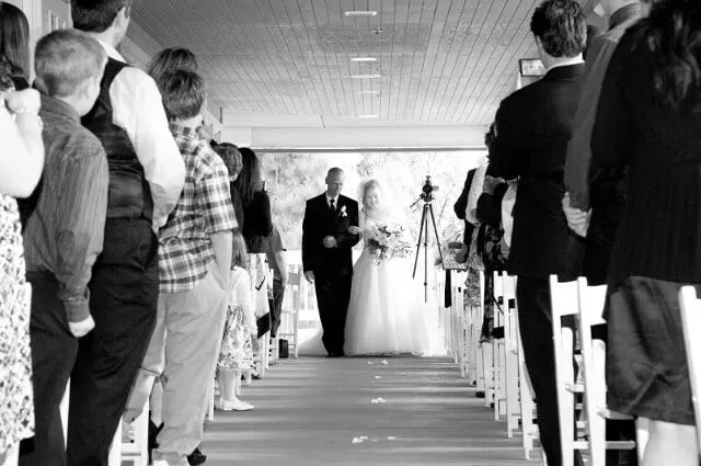 Black-and-white aisle view with guests on both sides and a groom walking toward the camera