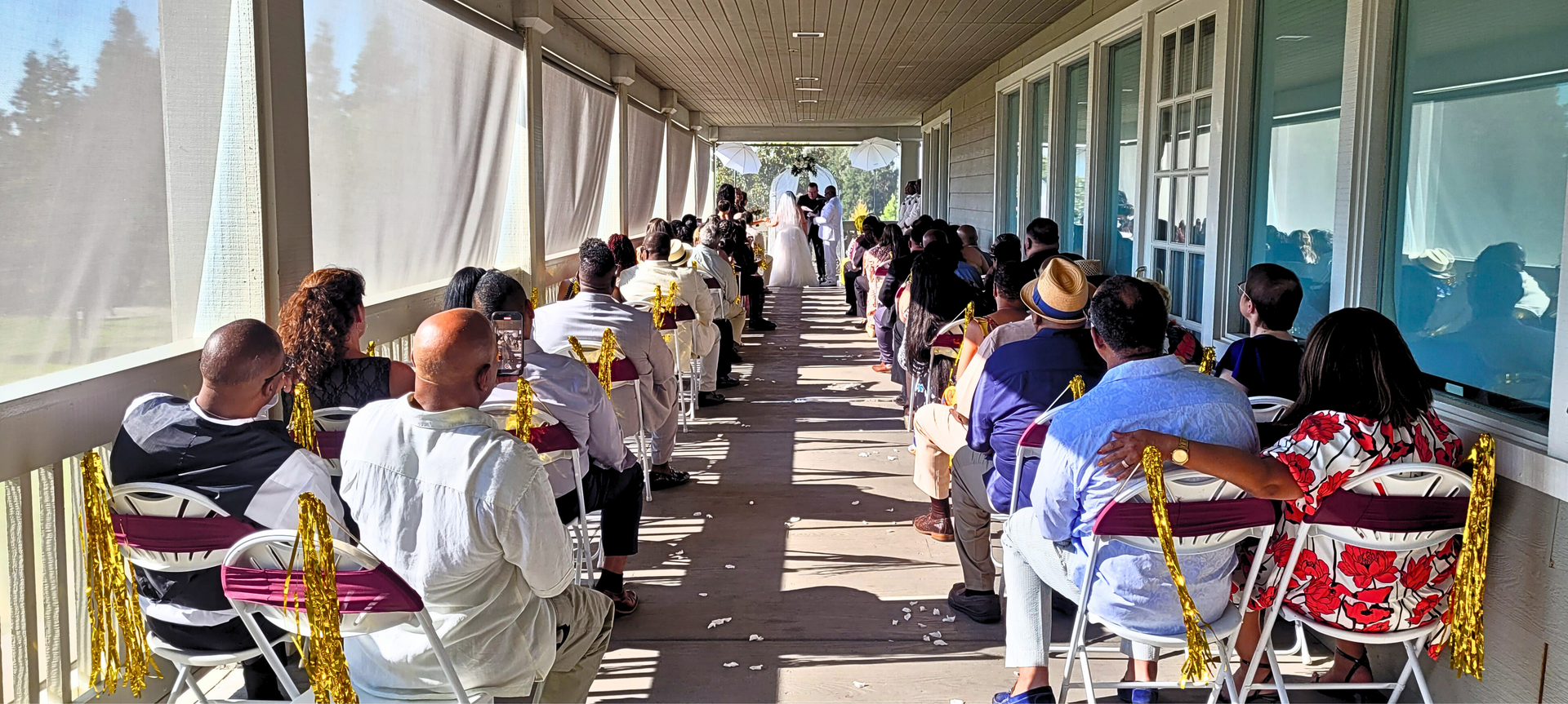 People seated in rows along a covered outdoor corridor, facing a speaker at the far end