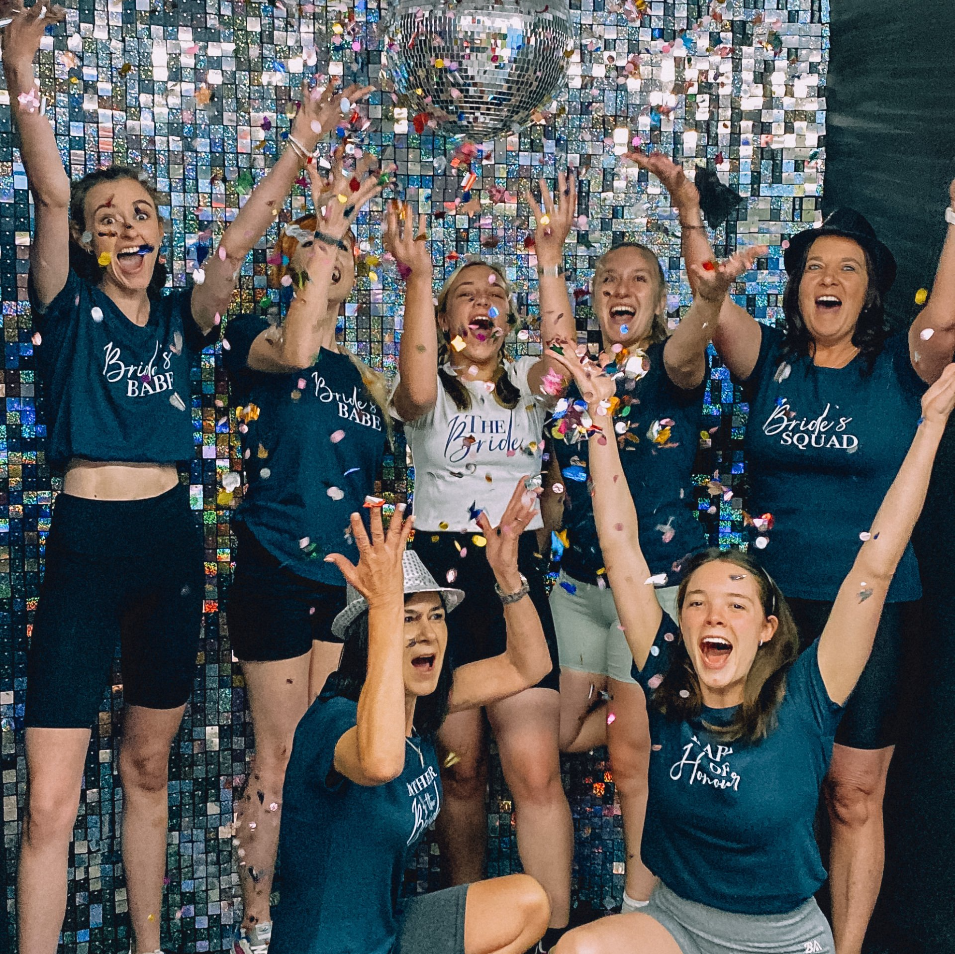 A cheerful group of young women posing together in sparkly outfits, smiling and celebrating with confetti falling around them during a joyful event.