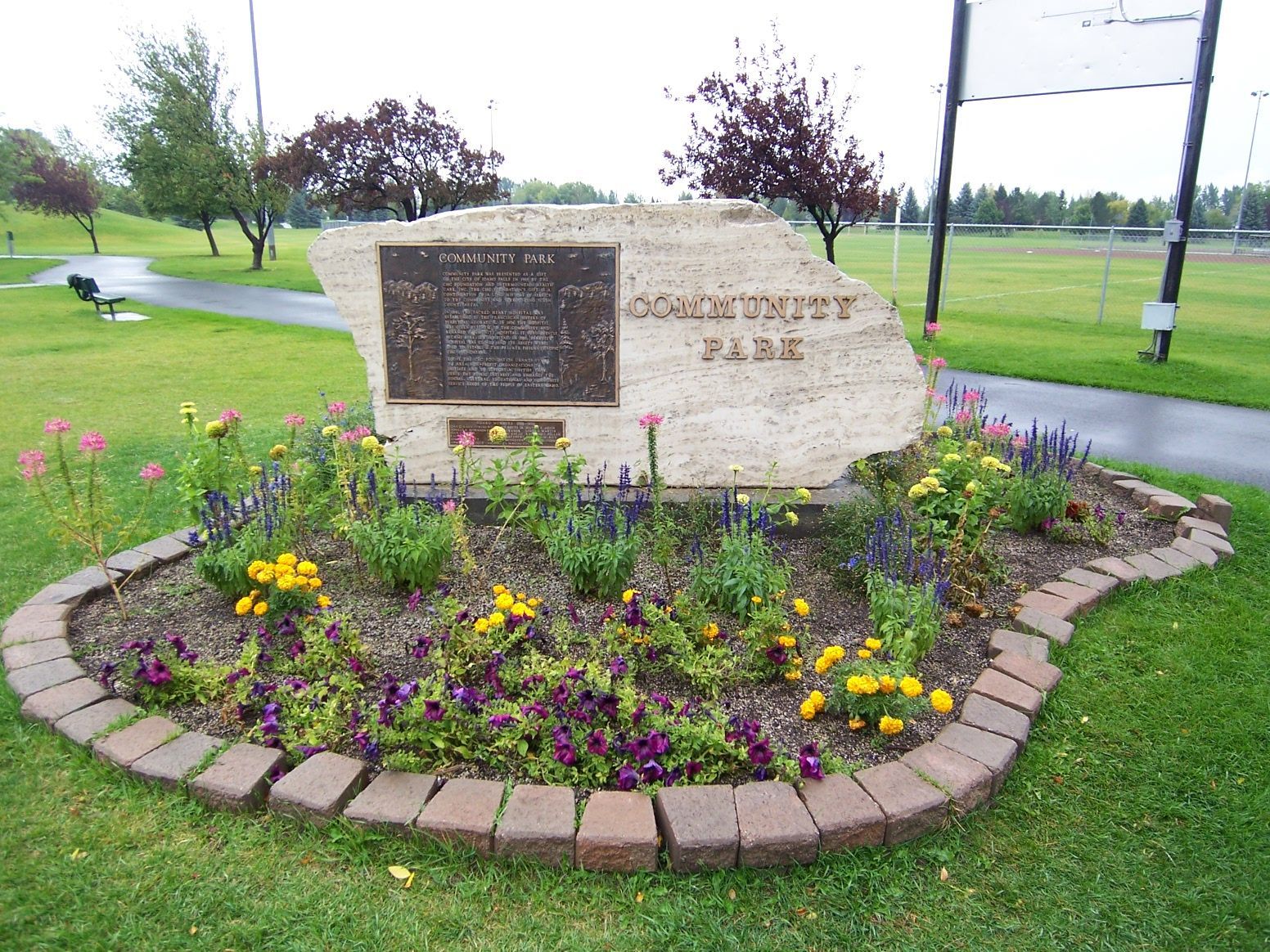 Community Park monument with plaque, surrounded by flowers and a brick border, set in a green park with trees.