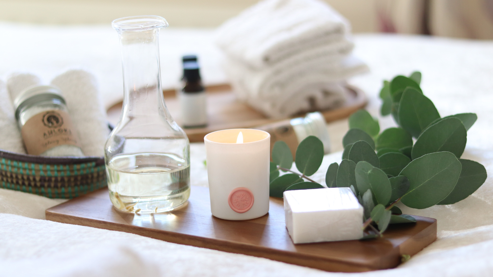 A spa-themed arrangement with a lit candle, oil decanter, eucalyptus sprig, bar of soap, and folded towels on a white bed.