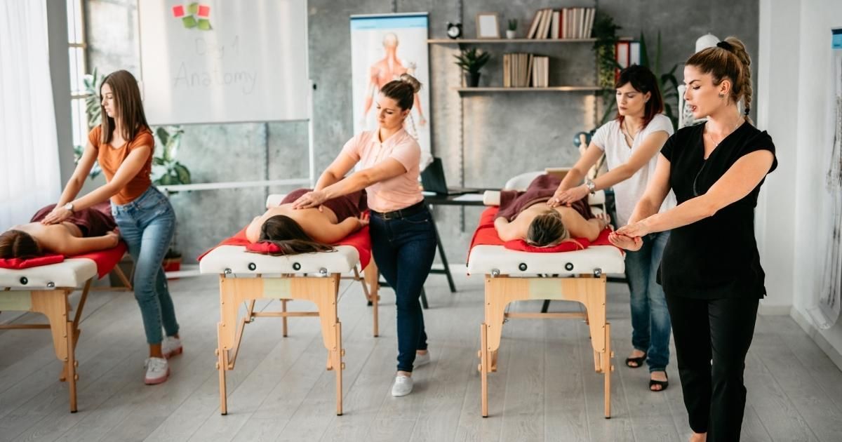 In a bright studio, three students practice massage techniques on people lying on tables, while an instructor observes.
