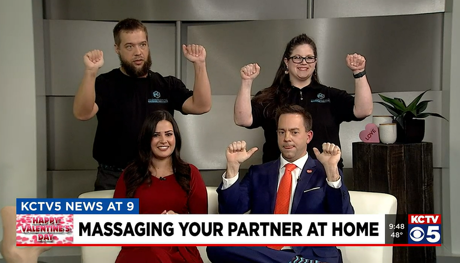 Four people on a news set pose with their hands raised, while a graphic onscreen reads, 
