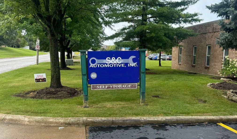 Sign for S&O Automotive, Inc., blue and white, with wrench graphic; in front of building, trees, and parking area | S&C Automotive Inc