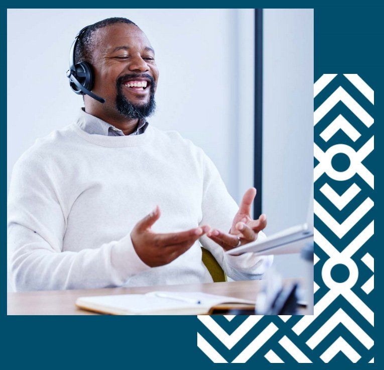 Smiling Black man wearing headset, gesturing while sitting at a desk. Office setting.