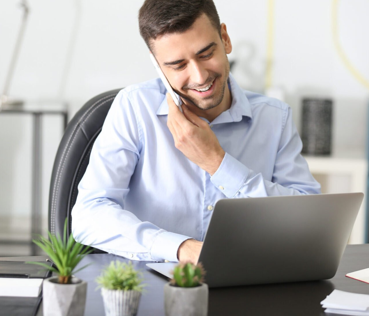 Man smiles while on the phone and works on a laptop in a light-filled office.