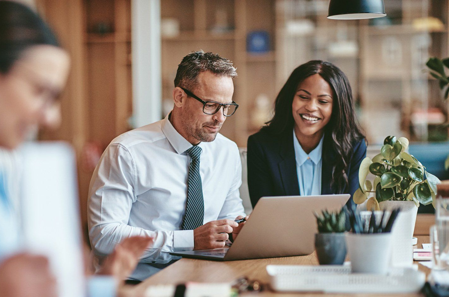 Coworkers collaborating at a laptop in a bright office, smiling over documents and a plant on the table