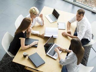 Team of five working around a table with laptops, papers, and a tablet in a modern office.