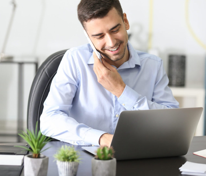 Man in a light blue shirt smiling at a laptop, touching his neck at a desk with small potted plants.