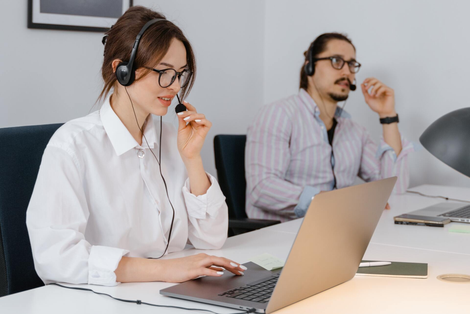 Woman in glasses wearing headset, using laptop; man in background, wearing headset.