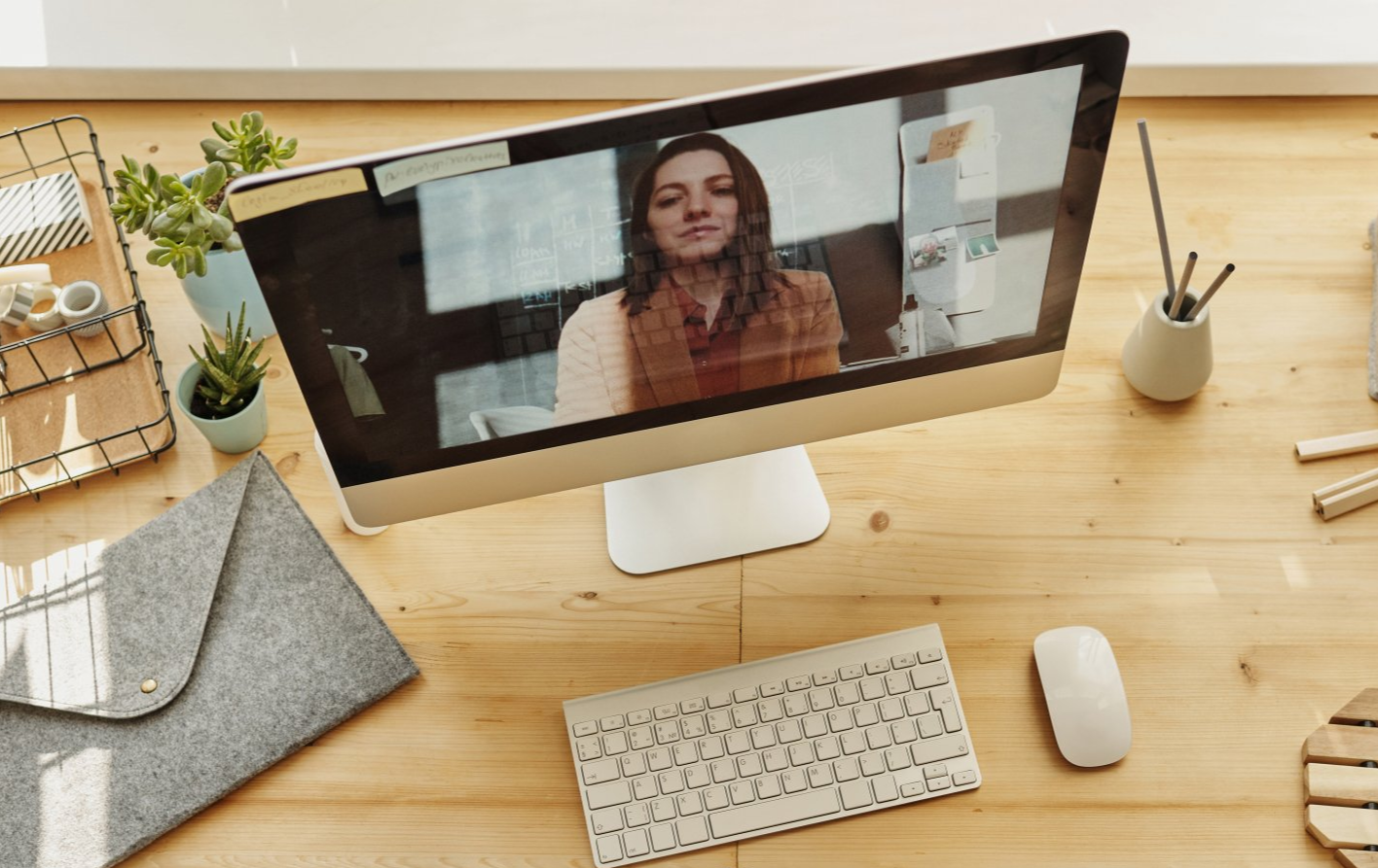 Person in a video call on a desktop monitor at a wooden desk with a notebook, coffee, and laptop nearby.