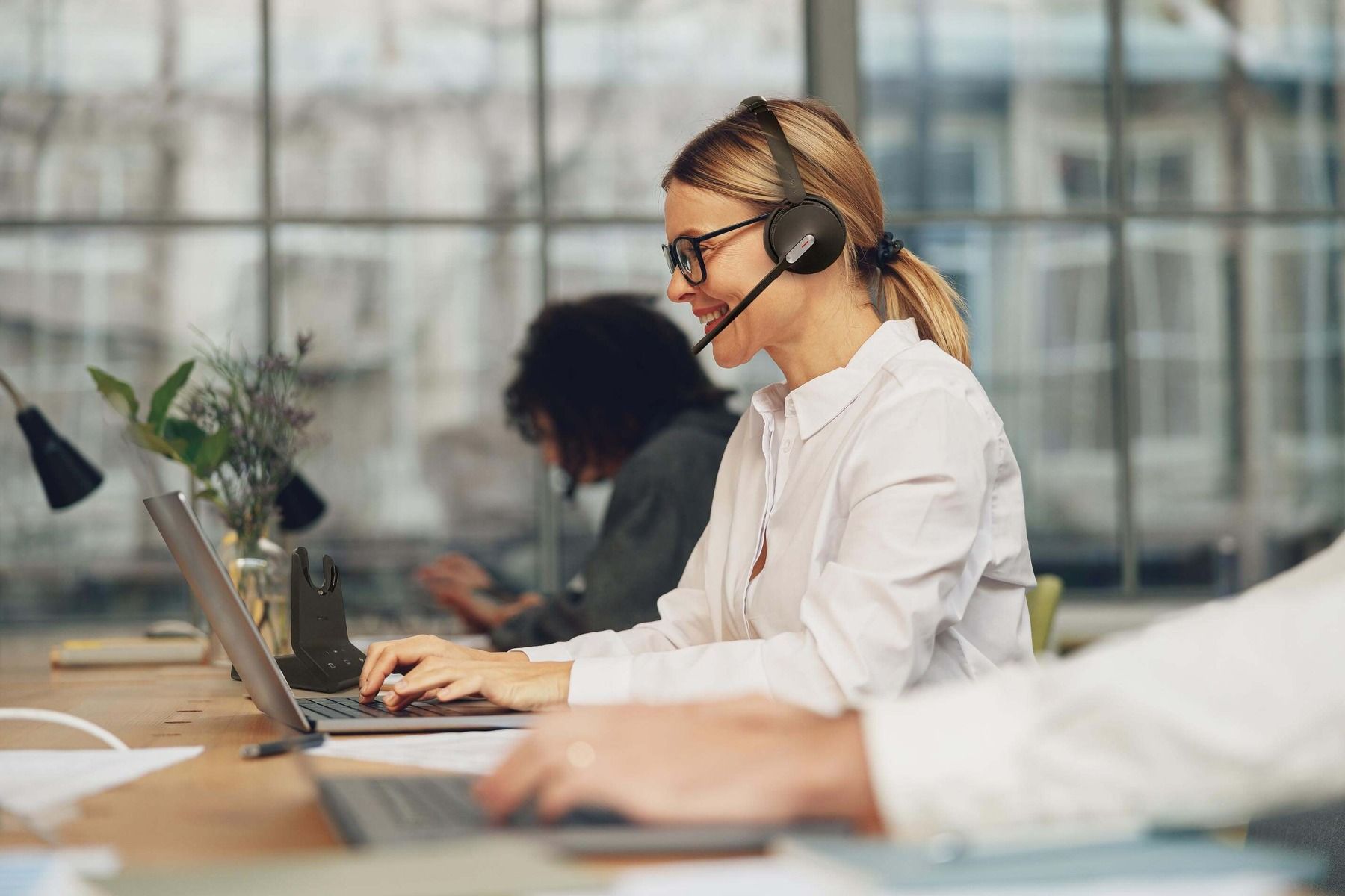 Office worker wearing a headset typing on a laptop at a desk in a bright workspace
