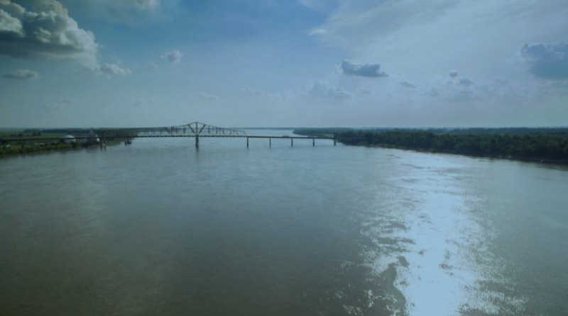 Wide river under cloudy sky with a long bridge in the distance.