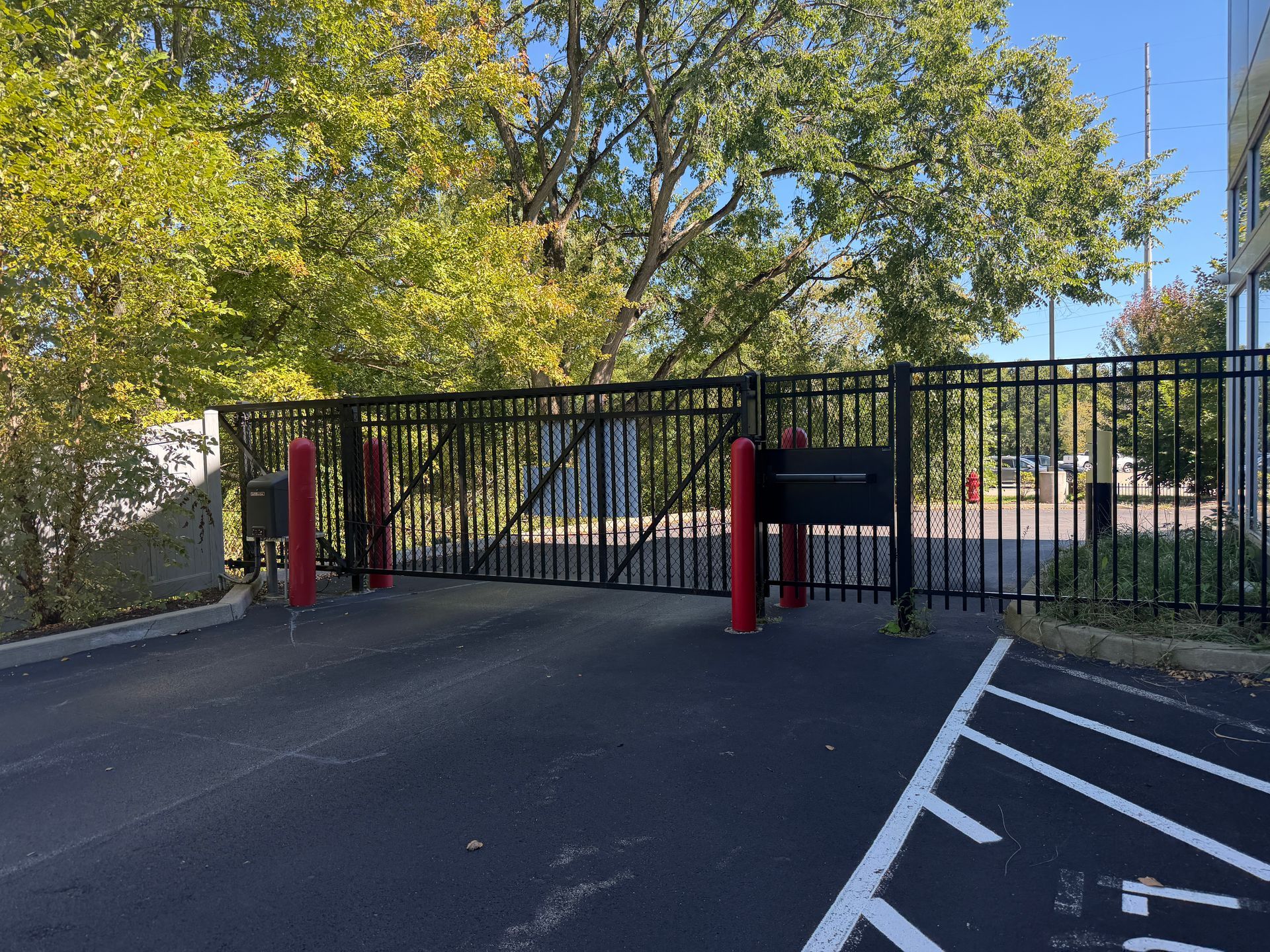Black metal gate blocking a driveway, with red posts and trees beside an asphalt road