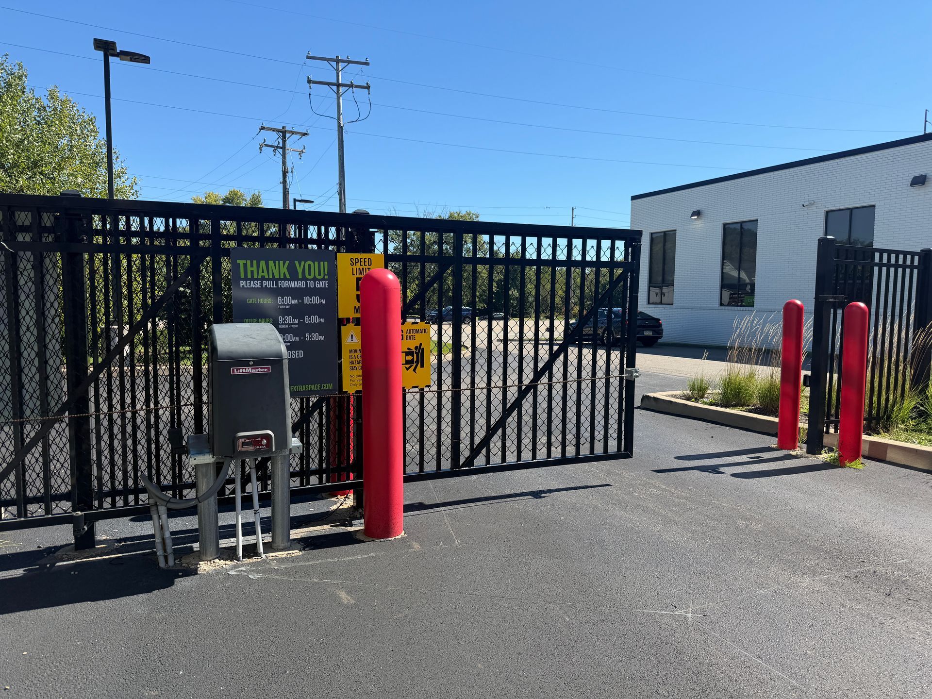Black metal gate with red bollards at a parking lot entrance beside a white industrial building.