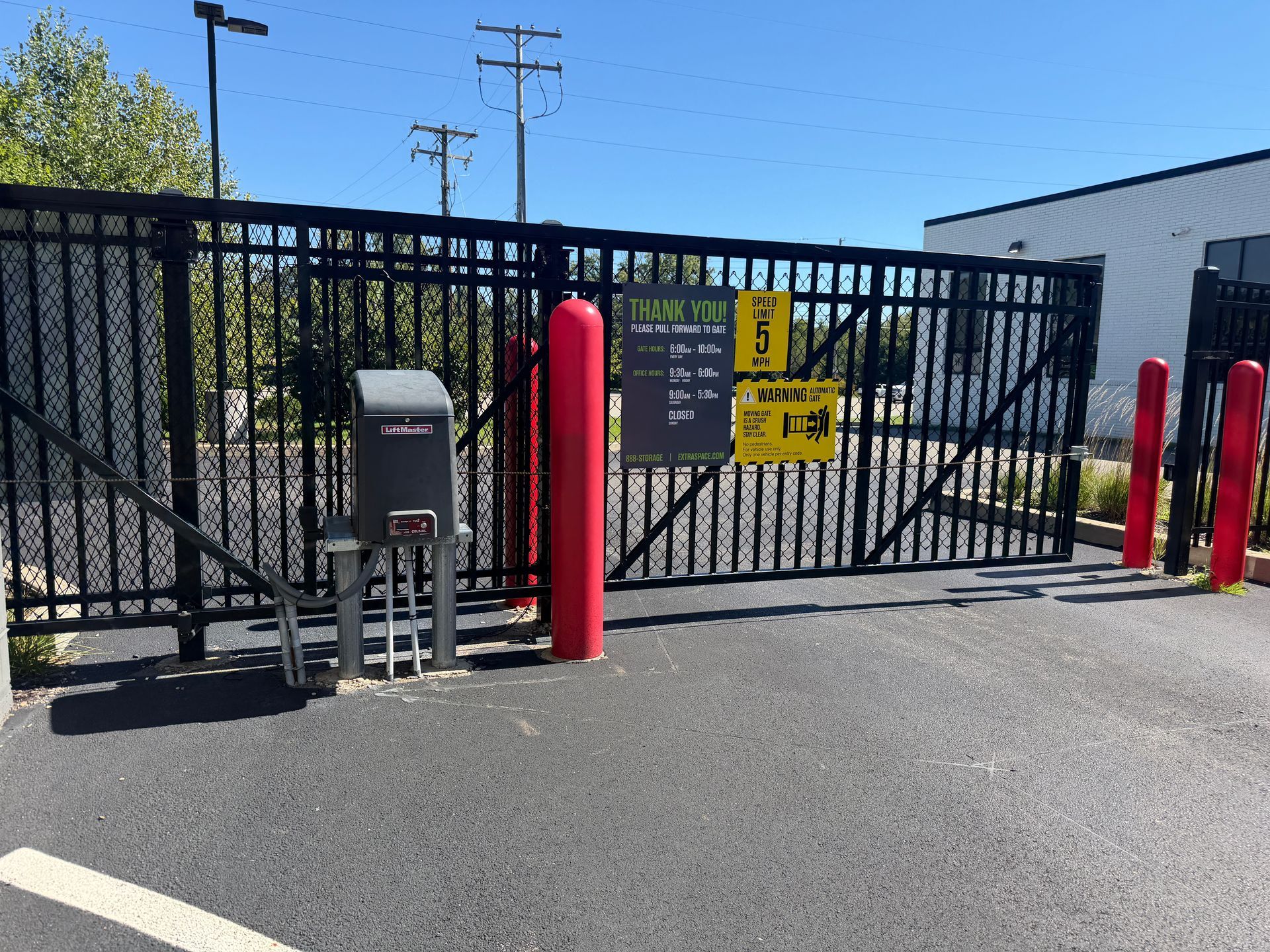Industrial gate with keypad access, warning sign, and red bollards beside a fenced lot