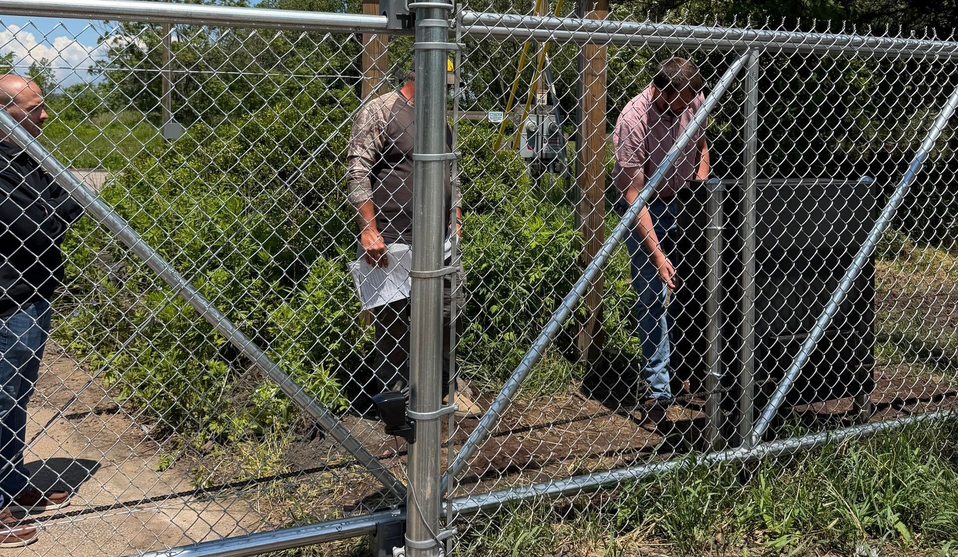 People behind a chain-link fence beside a sign and gate on a sunny day.