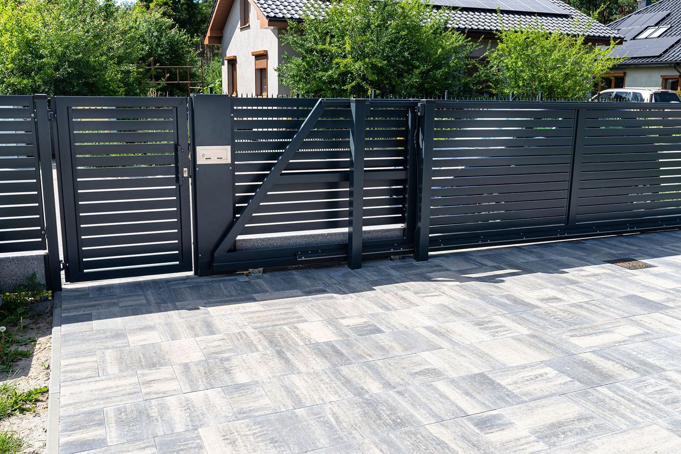 Metal gate, arched top, vertical bars, in front of a house with bushes on either side.