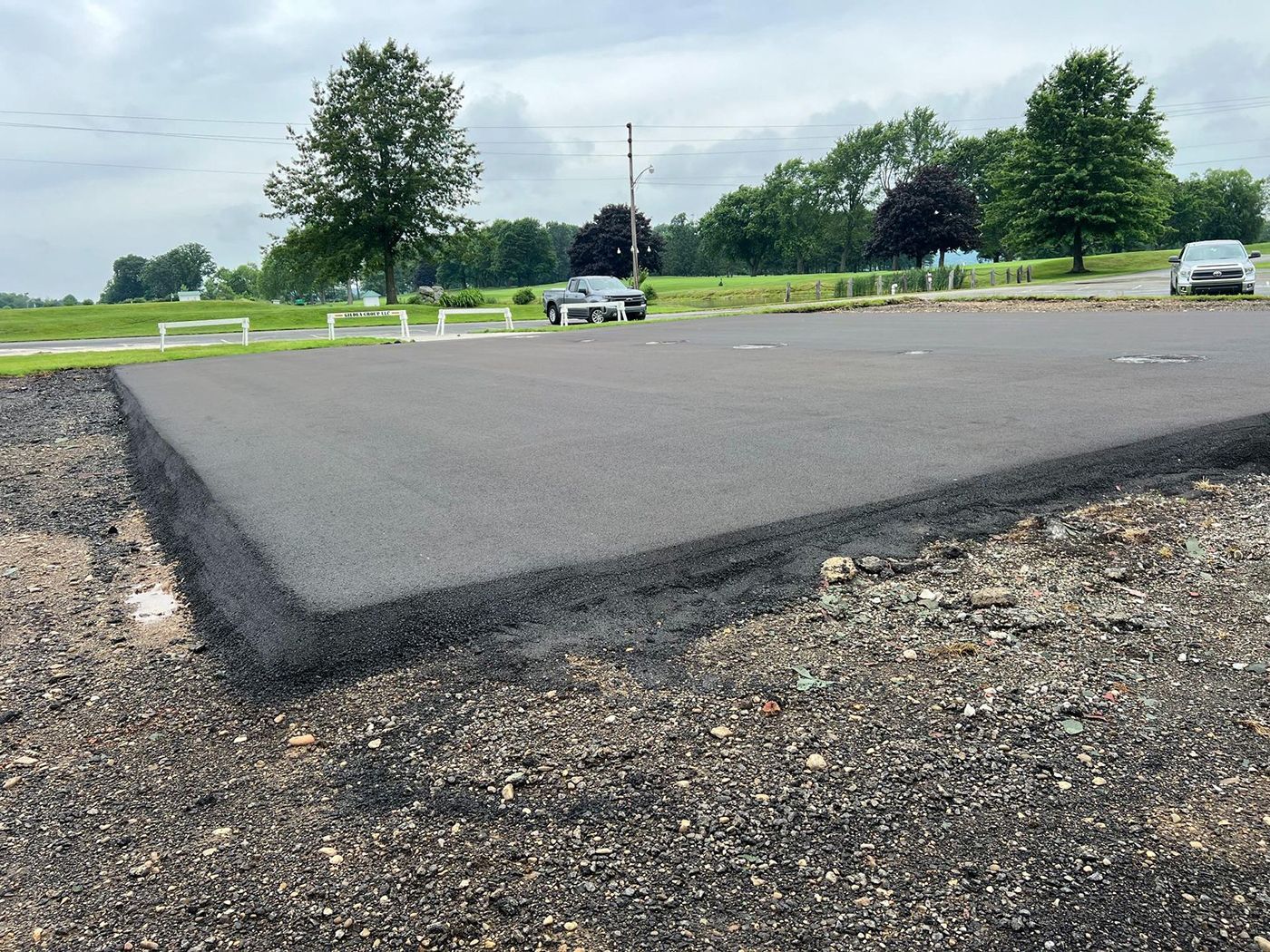 Low-angle view of a paved road edge with gravel shoulder, white barriers, and trees under a cloudy sky.