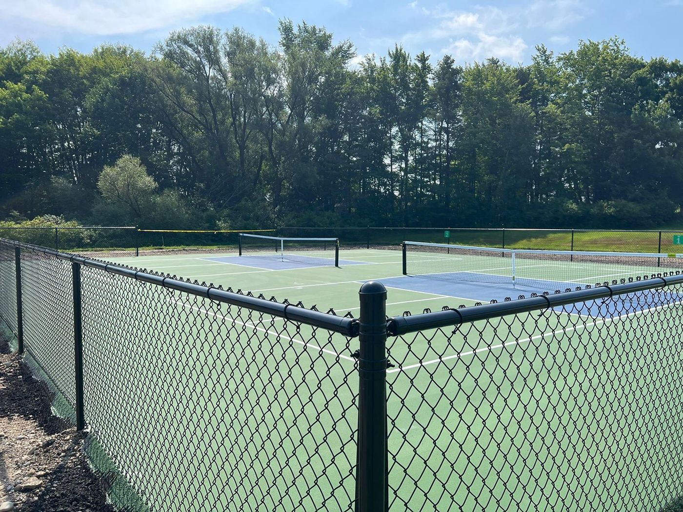 Tennis court behind a chain-link fence, with trees and blue sky in the background.
