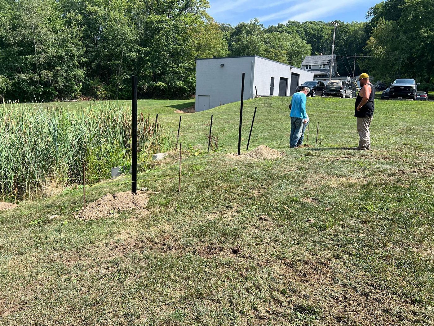 Two people standing by a fenced garden next to a white building on a sunny grassy field
