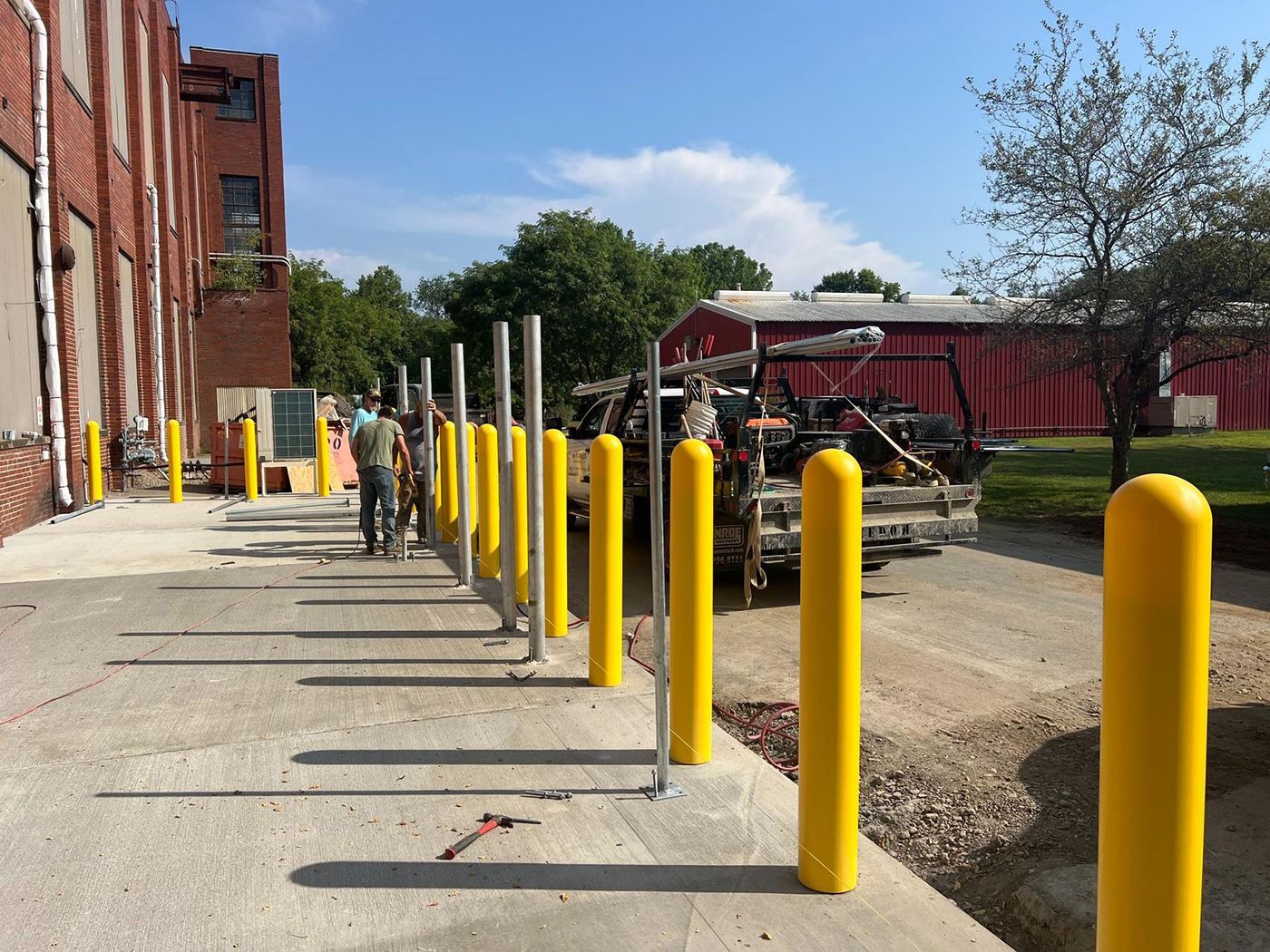 Concrete walkway beside brick building with yellow bollards and workers near a utility truck and trailer.