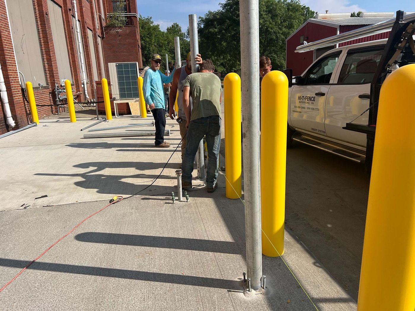 Pedestrians walking along a sidewalk beside a building, with yellow bollards and parked cars nearby.