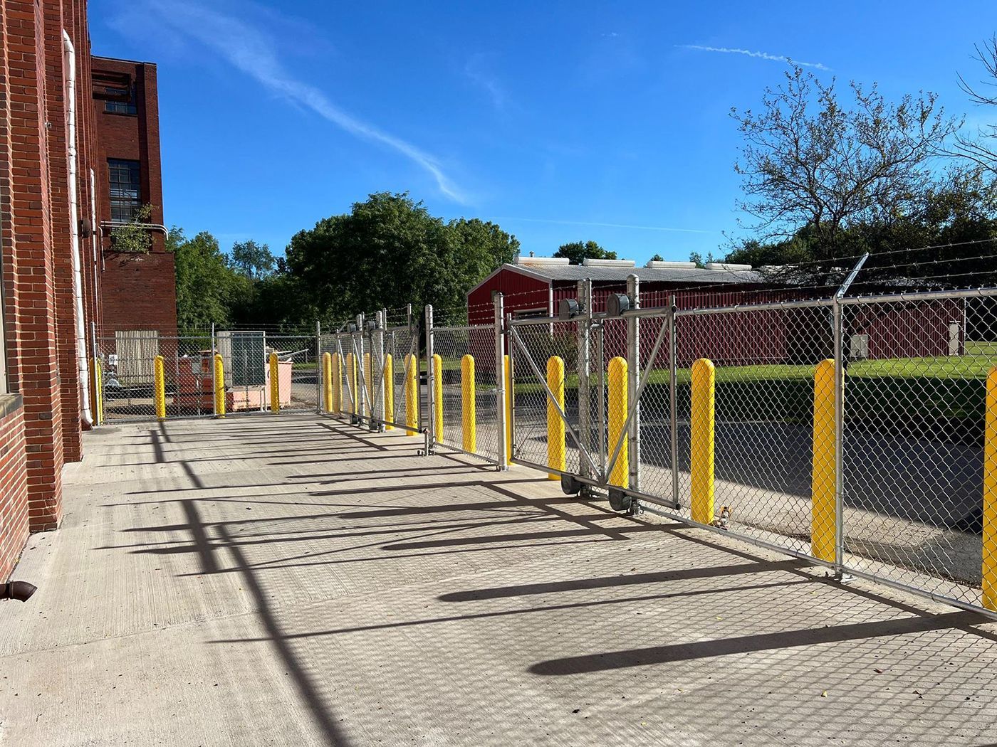Sunlit alley beside a brick building, lined with yellow bollards and chain-link fencing under a blue sky