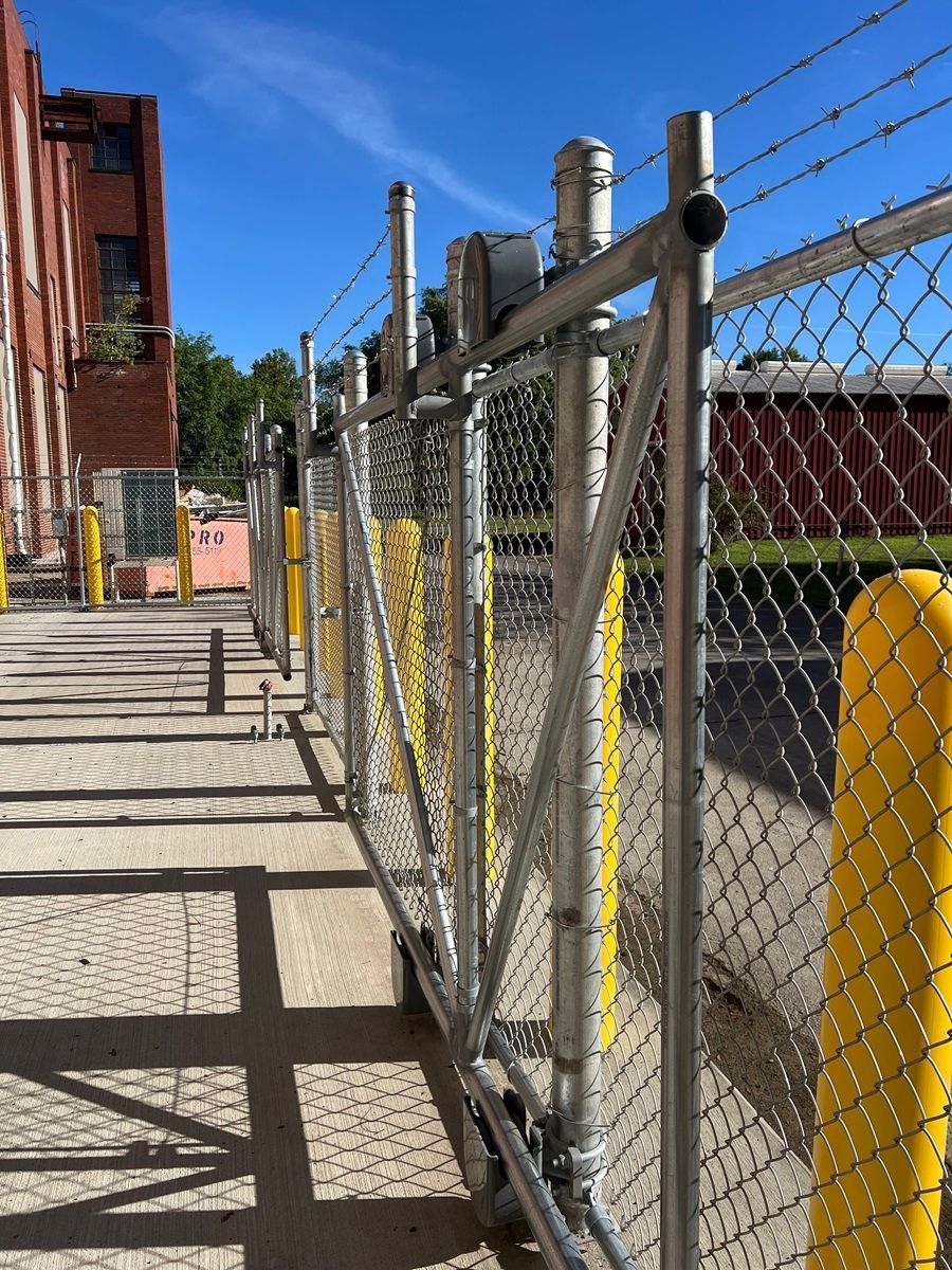 Chain-link fence along a sidewalk beside an industrial building under bright sunlight.