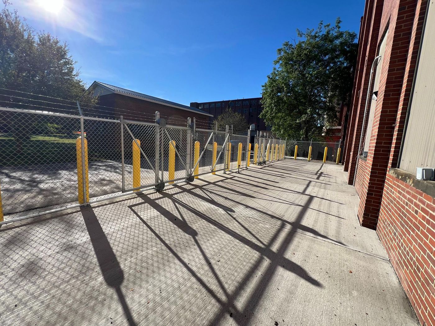 Sunlit sidewalk beside a brick building and chain-link fence, lined with yellow bollards and long shadows