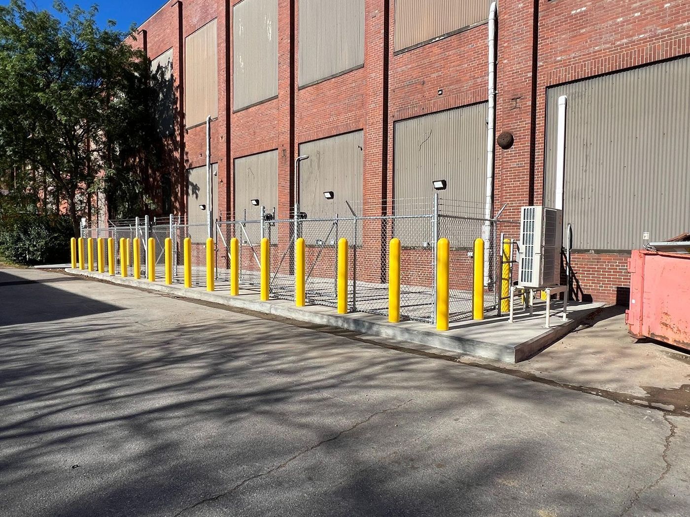 Yellow bollards line a sidewalk beside a red brick industrial building with closed gray doors.