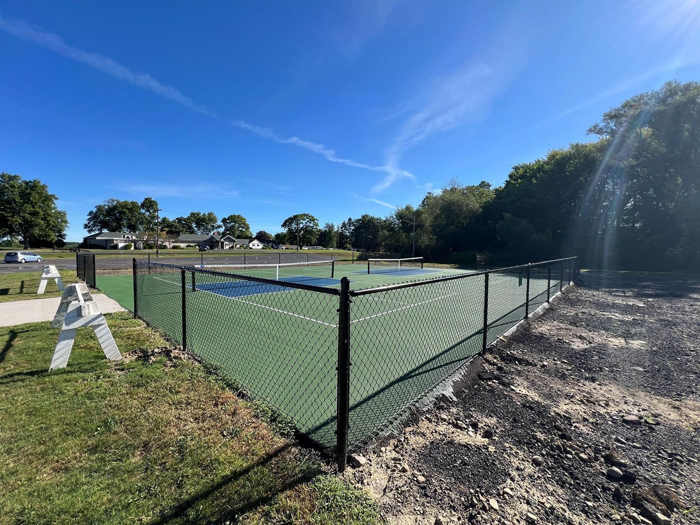 Chain-link fenced outdoor sports court under a bright blue sky, with grass, trees, and a dirt path nearby.